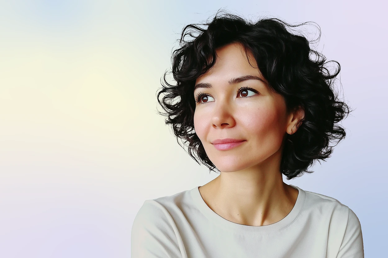 A woman with short, curly dark hair and a light complexion looks slightly to the side, smiling softly. She is wearing a light-colored shirt and is posed against a soft, pastel gradient background.