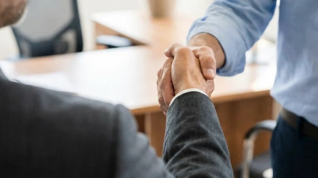 Two people in business attire shaking hands in an office setting, symbolizing agreement or partnership, with desks and chairs visible in the background.