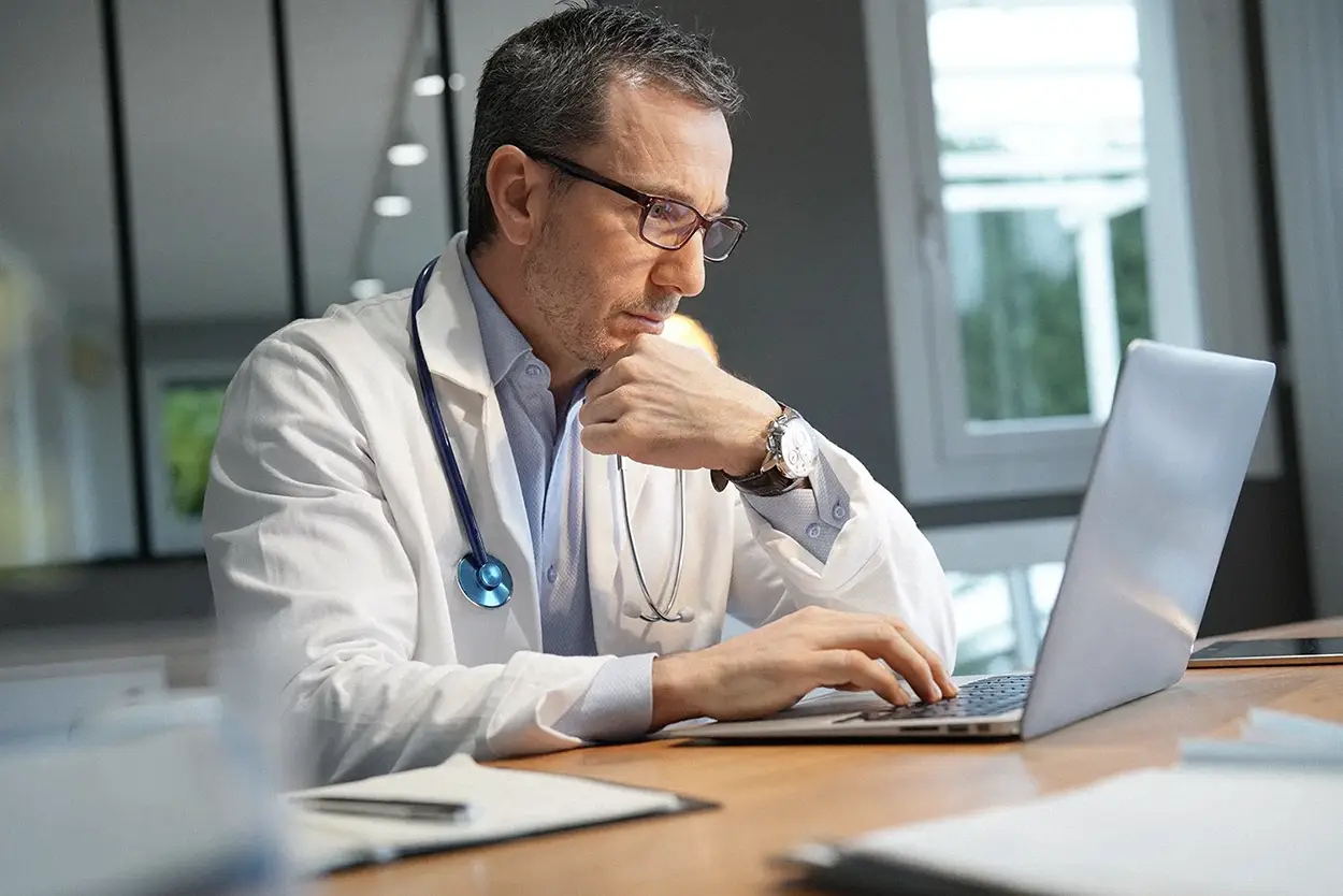 A doctor wearing a white coat and stethoscope sits at a desk, looking intently at a laptop screen with his hand resting on his chin.