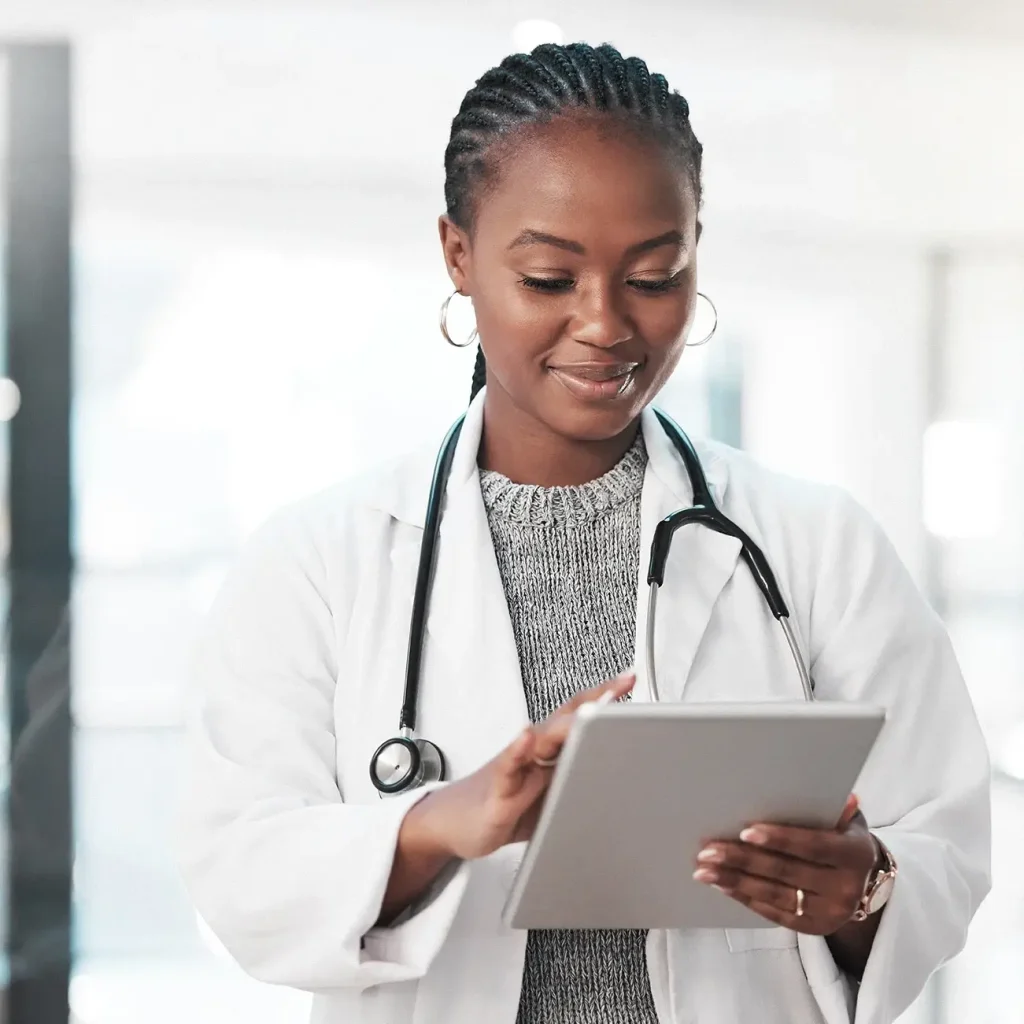 A doctor wearing a white coat and stethoscope smiles while using a digital tablet, standing in a bright, modern medical environment.