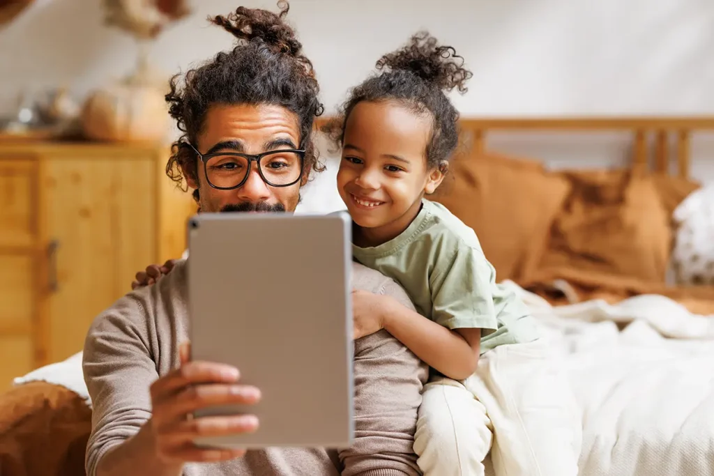 A man with glasses and a child smile while looking at a tablet together, sitting in a cozy bedroom with warm-toned decor.