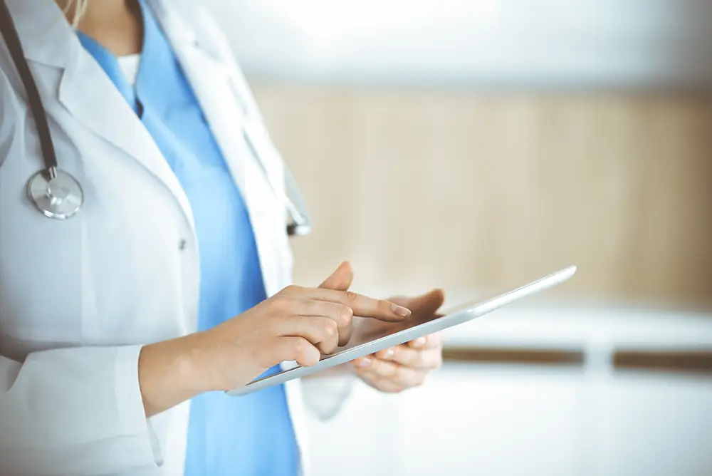 A medical professional wearing a white coat and stethoscope uses a digital tablet, standing in a bright, modern healthcare setting.