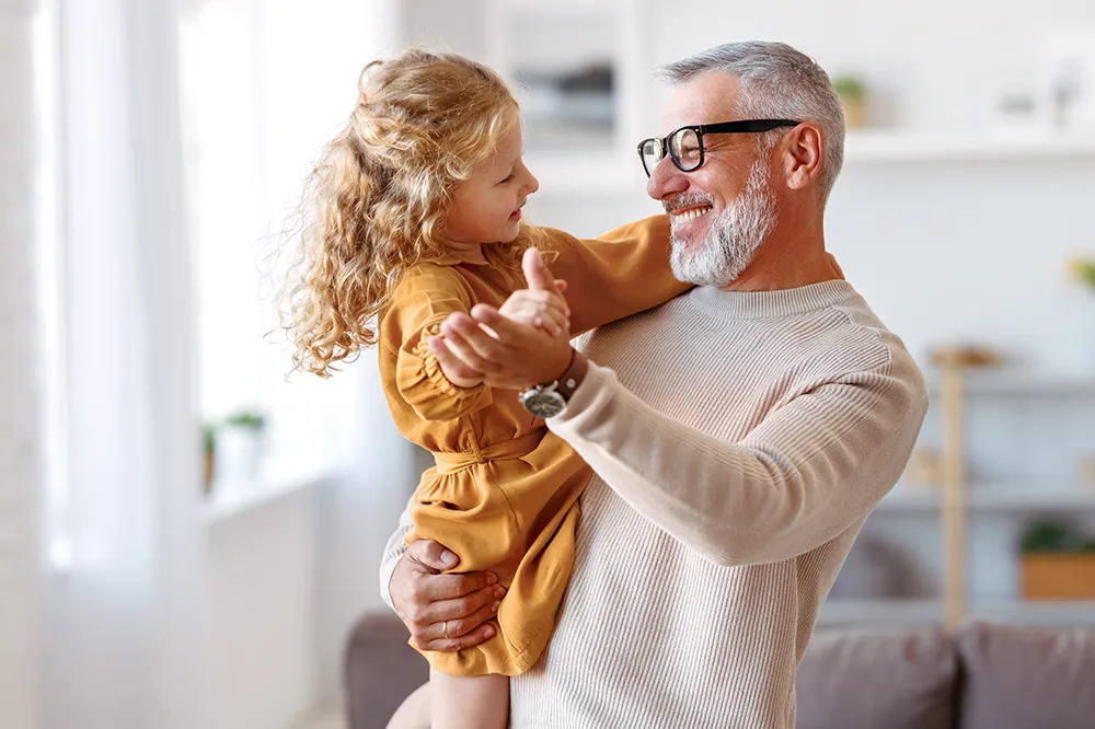 An older man with gray hair and glasses smiles while holding a young girl with curly blonde hair and a mustard dress. They look at each other happily in a bright living room.