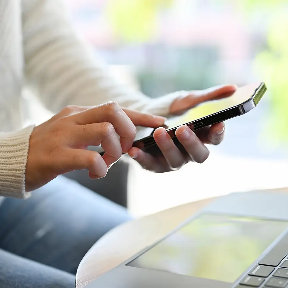 A person wearing a white sweater uses a smartphone with one hand while resting their other hand on their lap, seated next to an open laptop on a table.