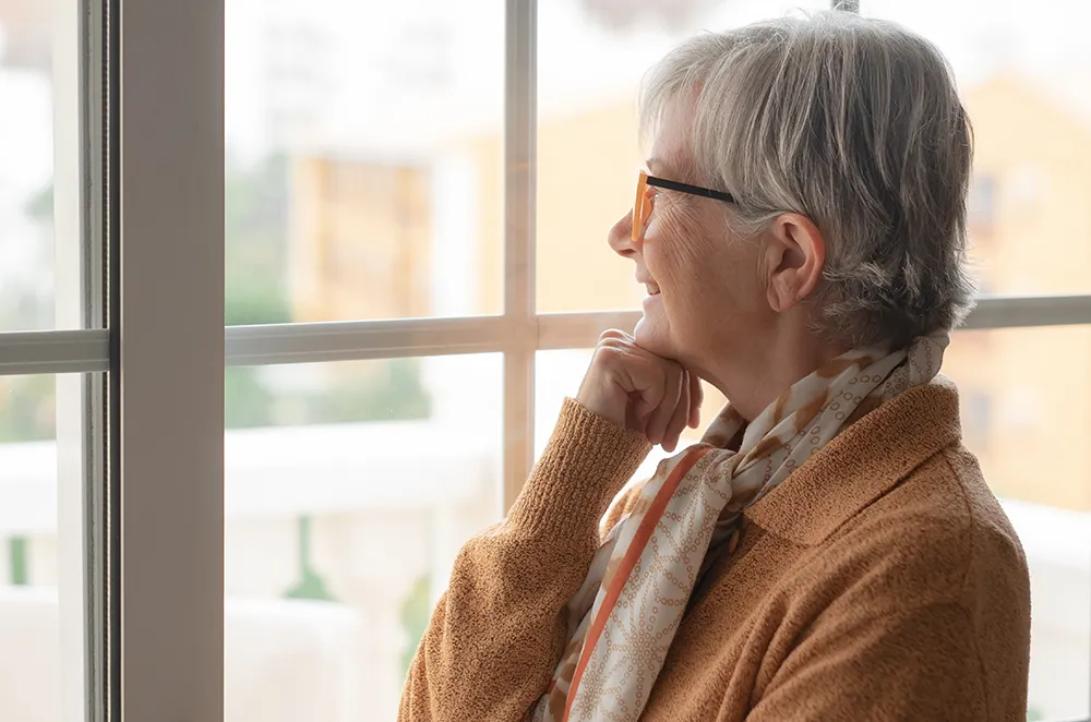 An elderly woman with short gray hair and glasses, wearing a light scarf and brown sweater, looks thoughtfully out a window with her chin resting on her hand.