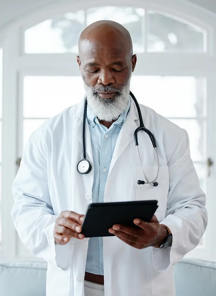 A doctor with a gray beard and stethoscope around his neck is wearing a white coat and looking down while using a digital tablet in a bright room.