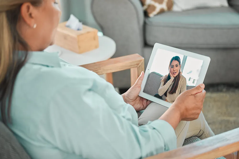 A woman sits in an armchair holding a tablet, having a video call with another woman who appears on the screen, speaking and smiling.