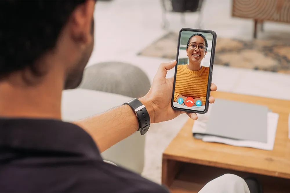 A person sits on a couch holding a smartphone, video chatting with a woman wearing glasses and a yellow sweater. The phone screen shows call controls, and a table with papers is in the background.