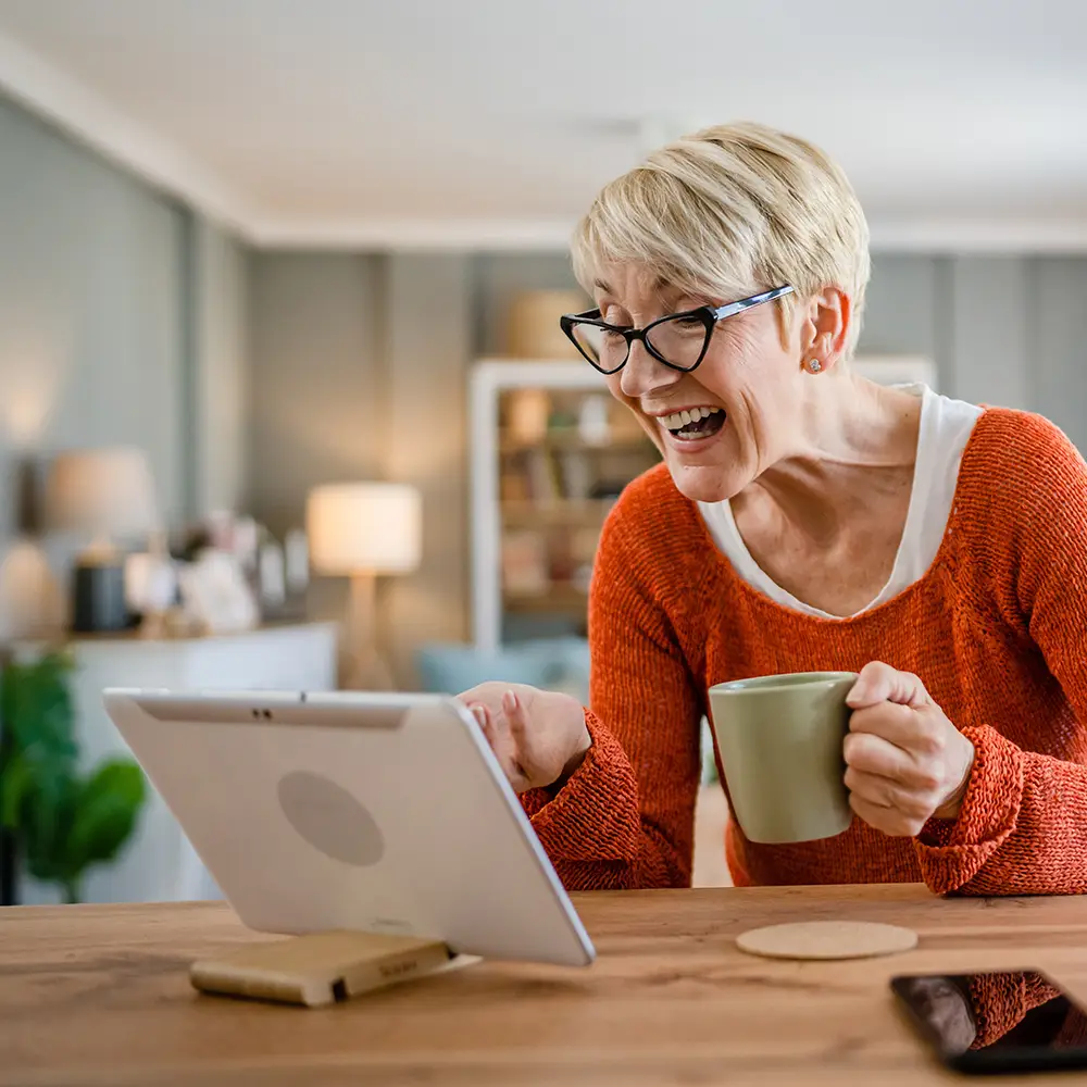 A smiling older woman with short blonde hair and glasses sits at a table, holding a mug and looking at a tablet screen. She appears to be having a video call in a cozy, well-lit living room.