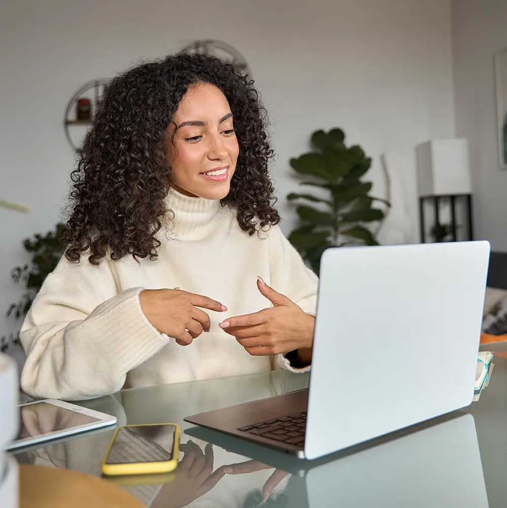 A woman with curly hair in a cream turtleneck sweater sits at a glass table, smiling and gesturing while video chatting on a laptop. A smartphone and notepad are beside her; a plant is in the background.