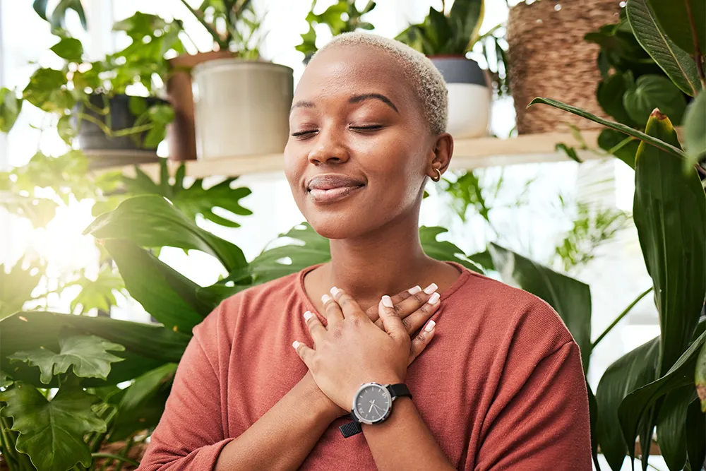 A person with short hair stands among green plants, eyes closed and hands crossed over their chest, appearing peaceful and content. They wear a watch and a rust-colored top. Sunlight filters through the background.