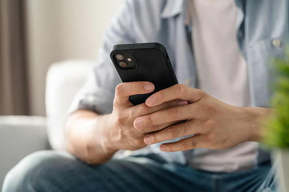 A person sits indoors, holding a black smartphone with both hands. The persons face is not visible, and they wear a blue shirt and jeans. The background is softly blurred, suggesting a home setting.