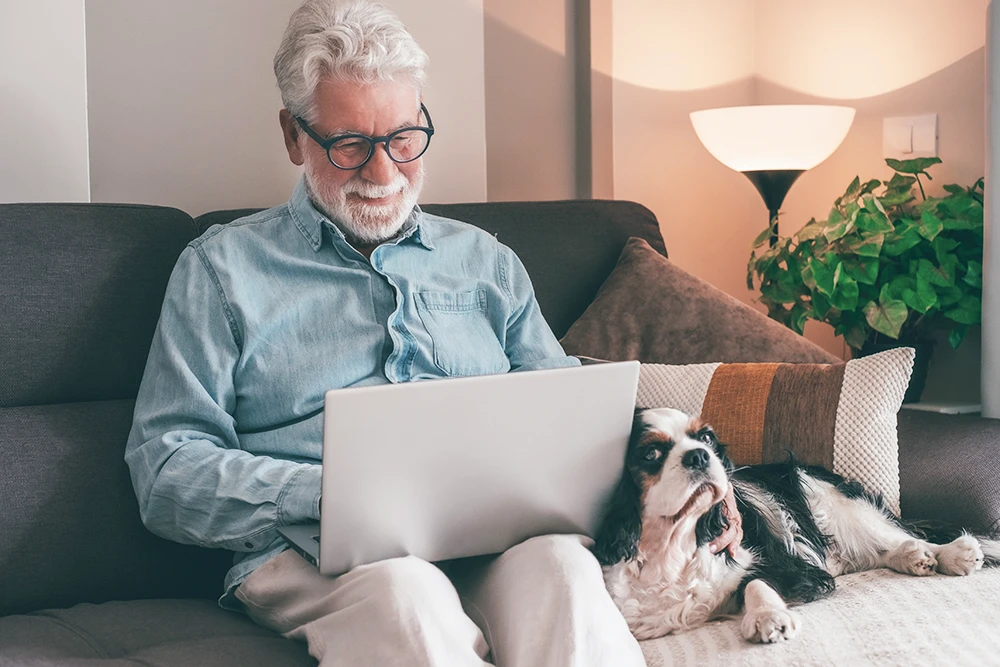 An older man with gray hair and glasses sits on a sofa, smiling while using a laptop. A black and white dog is lying beside him on the couch, and there is a lamp and plant in the background.