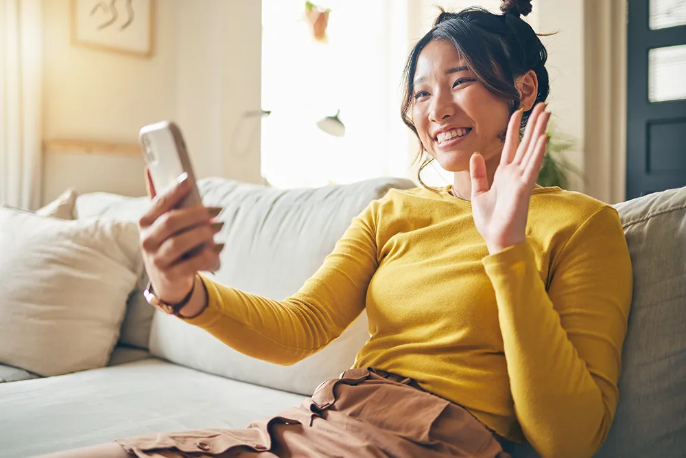 A young woman sitting on a couch, smiling and waving while holding a smartphone in her other hand, appearing to be on a video call in a brightly lit living room.