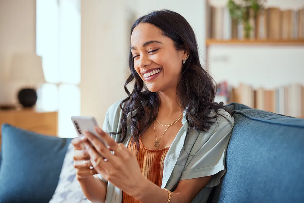 A smiling woman with long dark hair sits on a blue couch, looking at her smartphone. She wears a gold necklace, earrings, and a casual outfit, and appears happy and relaxed in a cozy, well-lit living room.