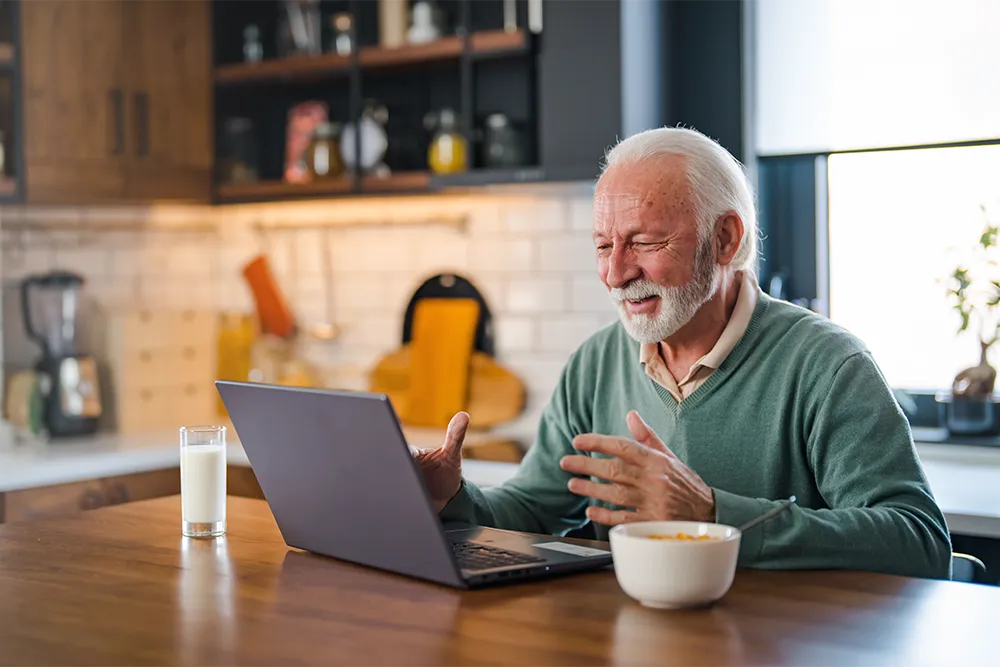 An elderly man with white hair and a beard is smiling and gesturing while video chatting on a laptop at a kitchen table, with a bowl of cereal and a glass of milk in front of him.