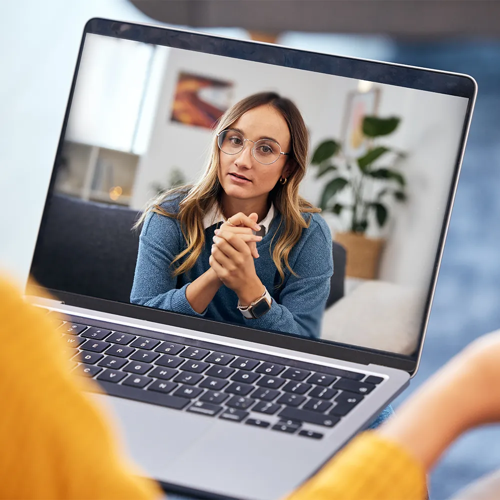 A woman with glasses, wearing a blue sweater, appears on a laptop screen during a video call, sitting indoors with a plant and framed picture in the background.