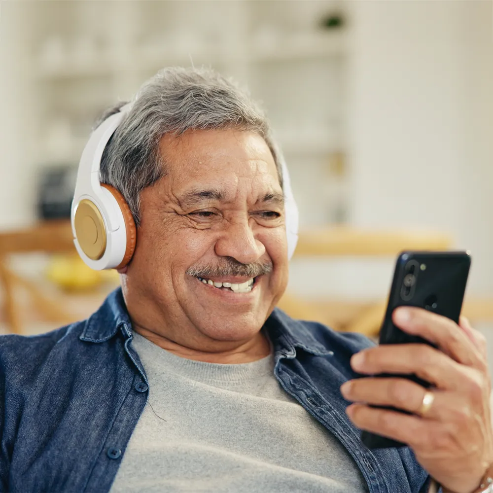 An older man with gray hair, wearing headphones and a denim shirt, smiles while looking at a smartphone in his hand. The background is softly blurred, suggesting a cozy indoor setting.