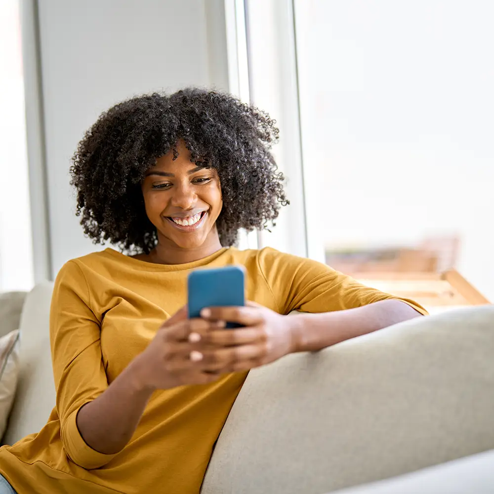 A woman with curly hair, wearing a mustard yellow shirt, sits on a couch and smiles while looking at her smartphone. Natural light fills the bright, modern room.