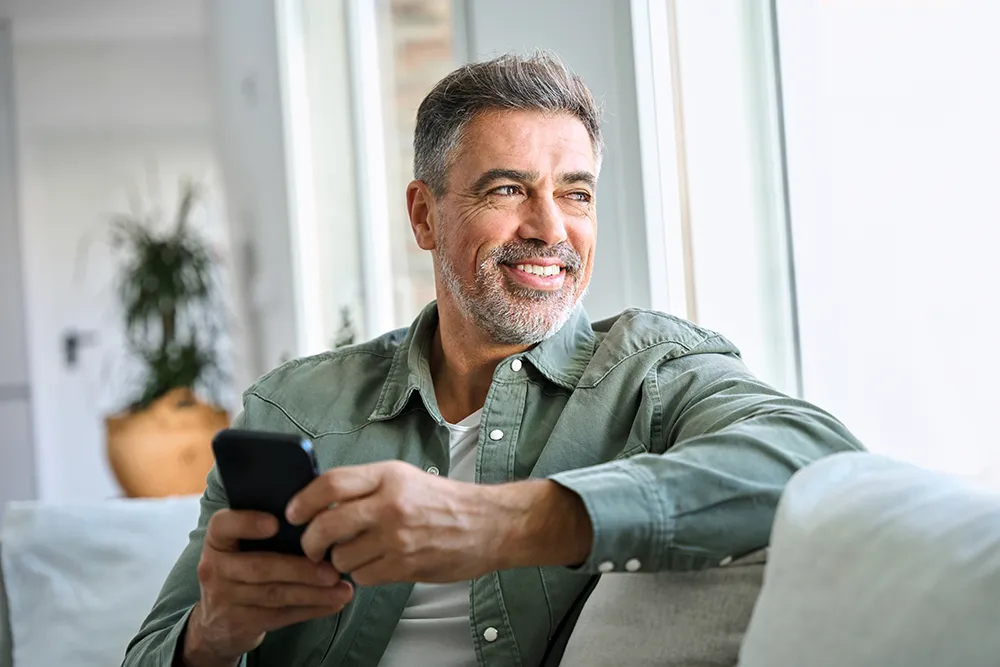 A middle-aged man with gray hair and a beard sits on a sofa, smiling and holding a smartphone. He wears a green button-up shirt and looks out a bright window, appearing relaxed and content.