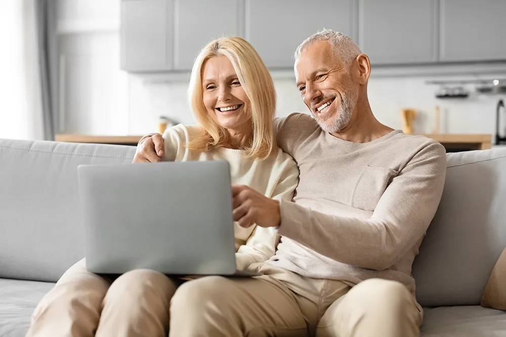 An older couple sits closely together on a couch, smiling and looking at a laptop screen, appearing happy and relaxed in a bright, modern living room.