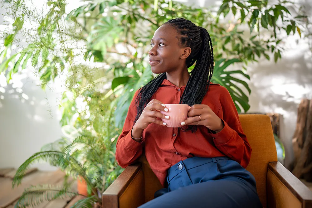 A woman with braided hair sits in an orange chair, holding a pink mug and smiling while looking to the side. She wears a rust-colored blouse and blue pants, surrounded by green indoor plants.