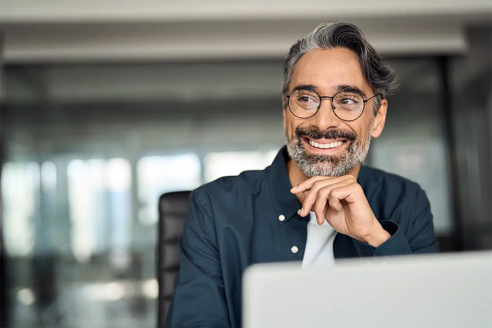 A smiling man with glasses and salt-and-pepper hair sits at a desk, resting his chin on his hand, looking to the side in a modern office setting.