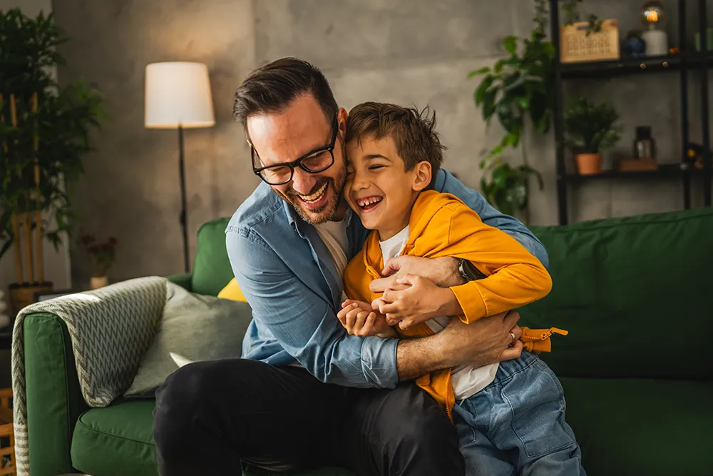 A bearded man wearing glasses hugs and laughs with a smiling young boy on a green couch in a cozy living room with plants and warm lighting.