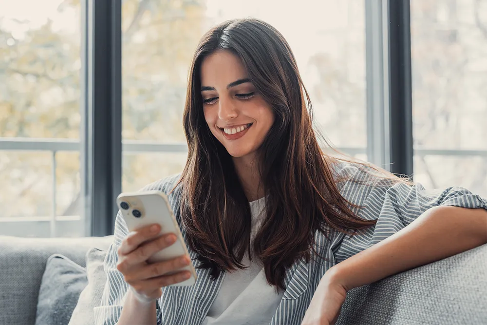 A young woman with long brown hair sits on a couch, smiling as she looks at her smartphone. She is wearing a striped shirt and is in a bright room with large windows in the background.