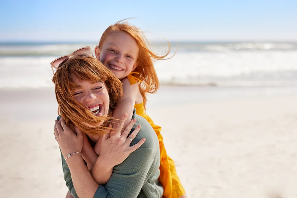 A smiling woman with red hair gives a piggyback ride to a laughing young girl in a yellow dress on a sunny beach, with waves and blue sky in the background.