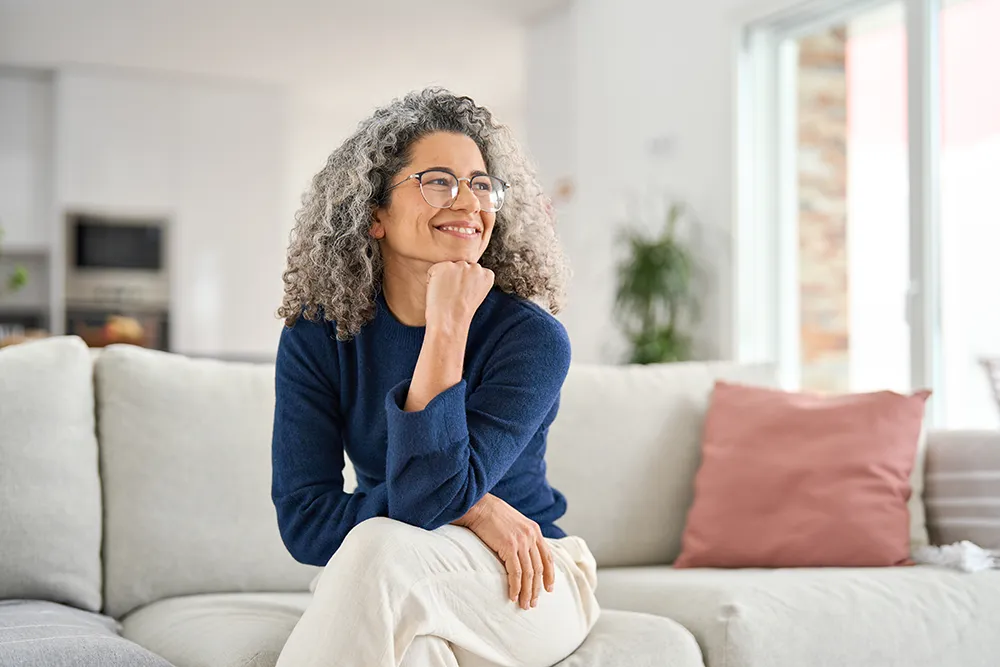Smiling older woman with curly gray hair and glasses sits on a light-colored sofa, resting her chin on her hand, wearing a blue sweater and beige pants in a bright, modern living room with natural light.