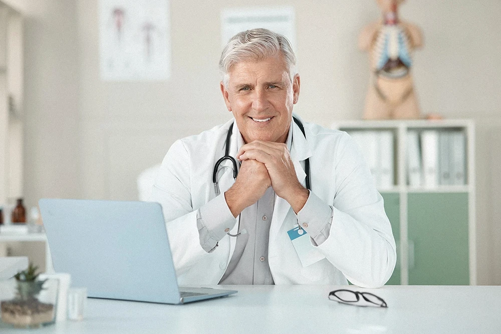 An older male doctor with gray hair and a stethoscope around his neck sits at a desk, smiling, with his hands clasped in front of a laptop in a bright medical office.