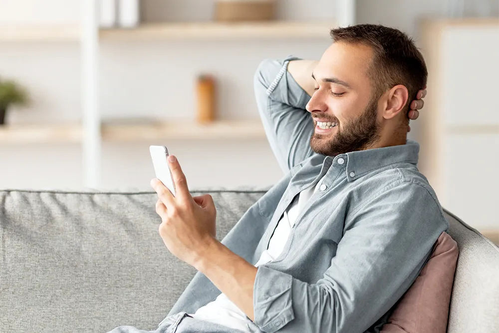 A smiling man sits on a couch, holding a smartphone in one hand and resting his other hand behind his head, appearing relaxed and happy while looking at the phone.