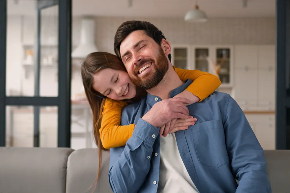 A smiling man sits on a couch while a young girl hugs him from behind, both looking happy and relaxed in a bright, modern kitchen setting.