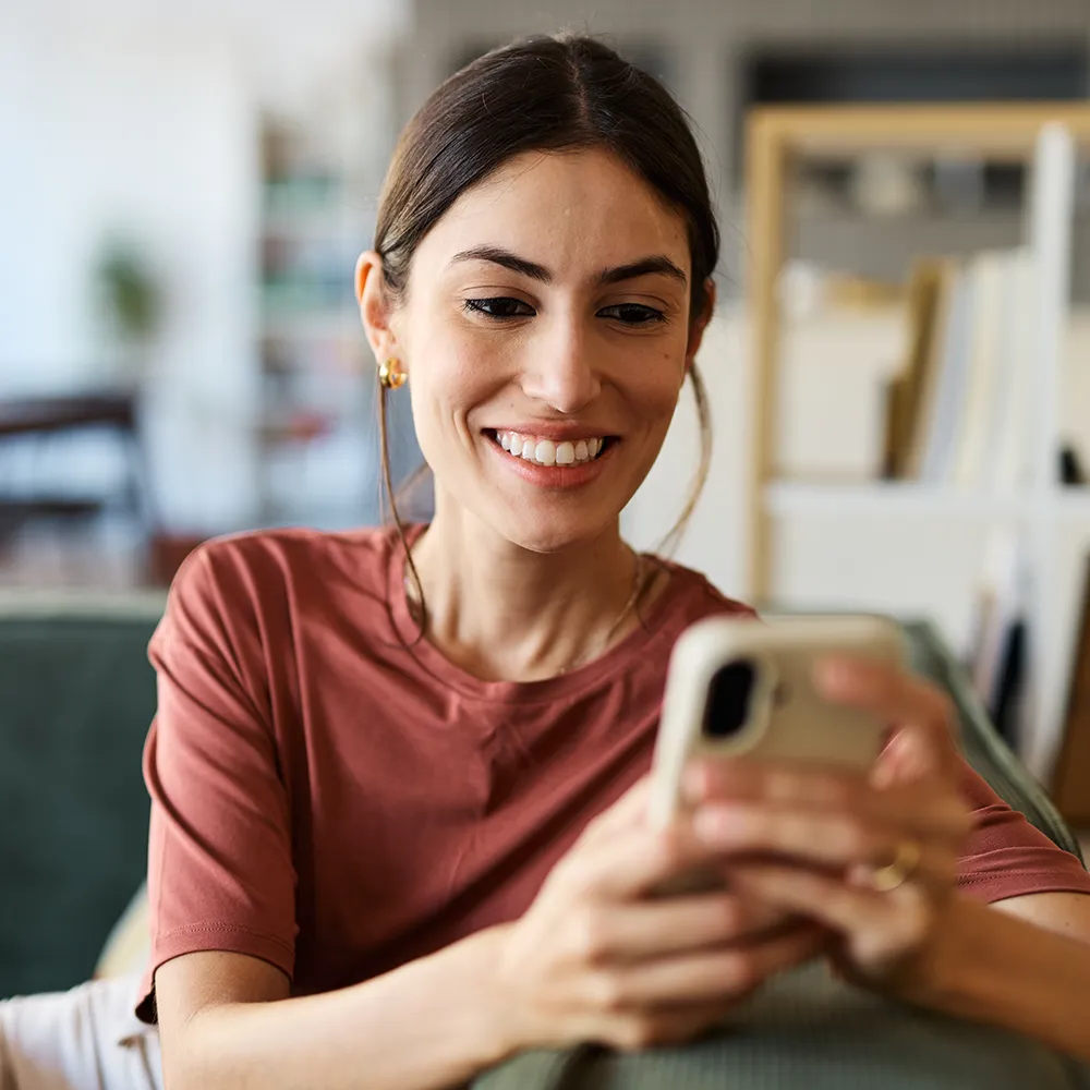 A woman with long brown hair, wearing a rust-colored shirt, sits on a couch smiling while looking at her smartphone. Bookshelves and a blurred background are visible behind her.