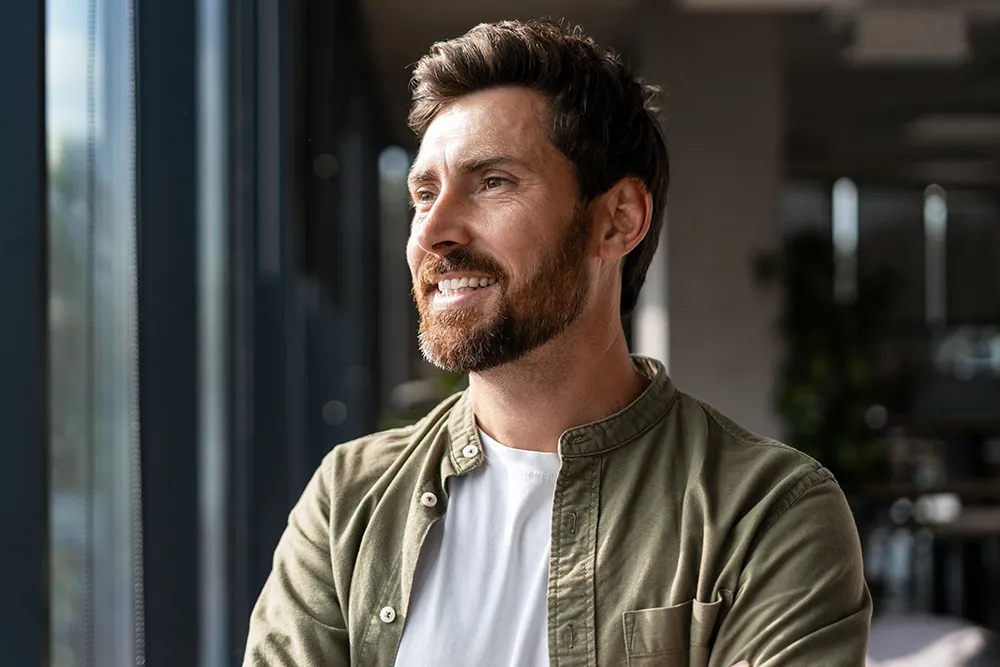 A bearded man with short brown hair, wearing a white t-shirt and an olive green button-up shirt, stands indoors and smiles while looking out of a window with his arms crossed.