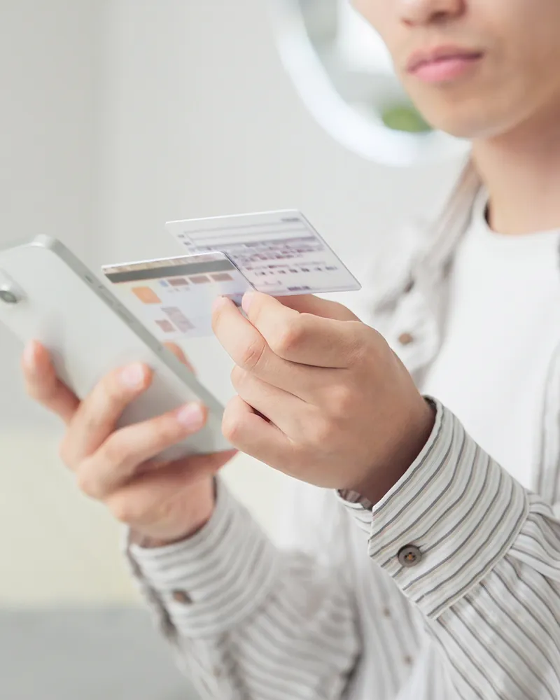 A person in a striped shirt holds a smartphone in one hand and a credit card in the other, appearing to make an online payment or purchase.