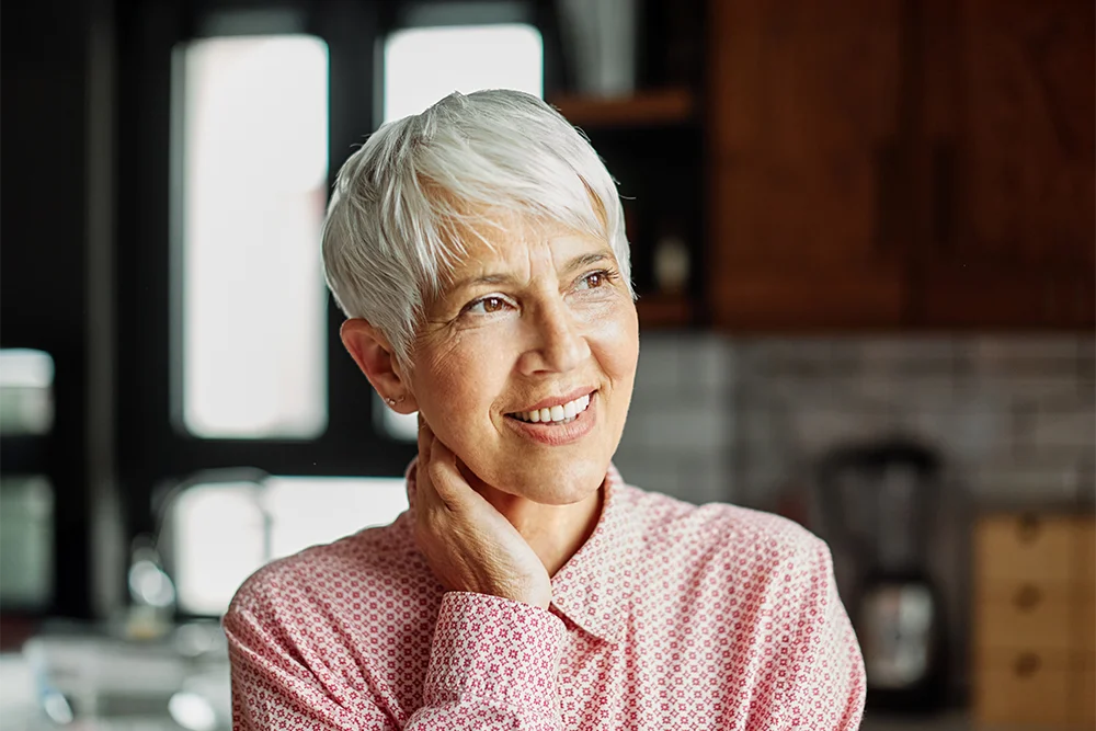 An older woman with short gray hair and a pink patterned shirt smiles, looking off to the side, with her hand gently touching her neck in a warmly lit kitchen.