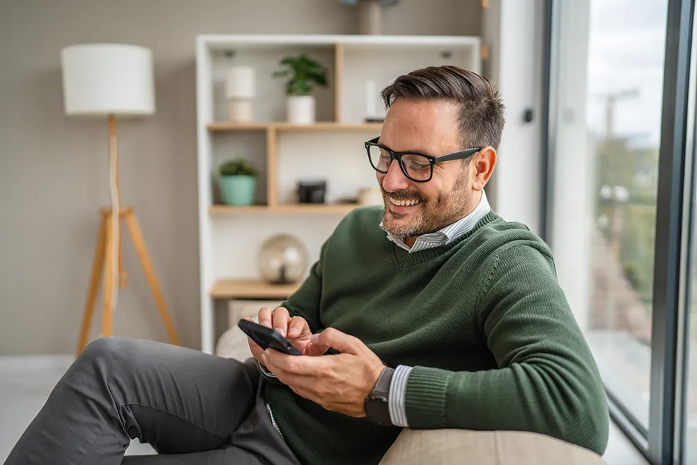 A man with glasses, wearing a green sweater, sits on a couch and smiles while using a smartphone. He is in a bright, modern living room with a large window and shelves in the background.