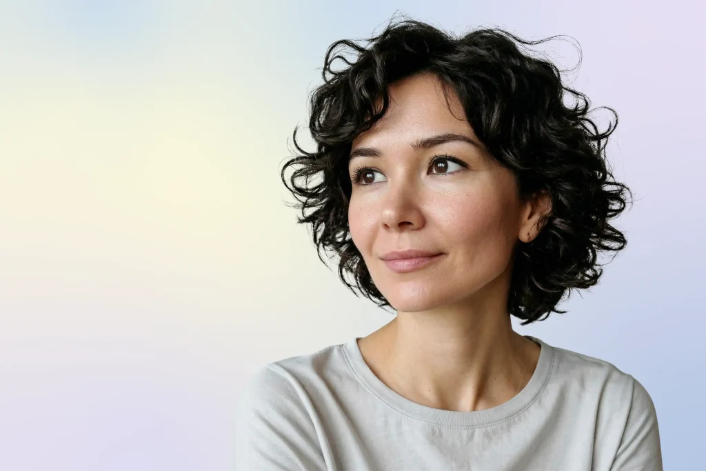 A woman with short, curly dark hair and a light complexion looks slightly to the side, smiling softly. She is wearing a light-colored shirt and is posed against a soft, pastel gradient background.